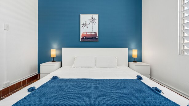 A bedroom with a blue wall, a white bed, two bedside tables with lamps, and a poster of a van and palm trees. - Otium Rentals San Juan City Apartment w Basketball Arcade (San Juan)