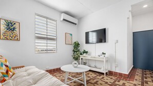 A modern living room with a white sofa, a small round table, a TV mounted on the wall, and a window with blinds. - Otium Rentals San Juan City Apartment w Basketball Arcade (San Juan)
