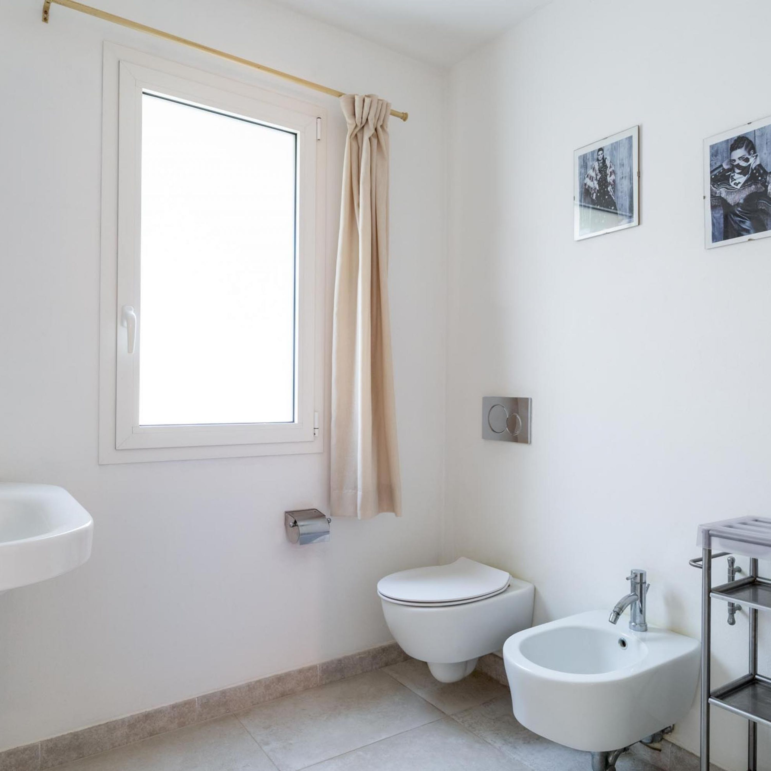 A bathroom with a toilet, bidet, sink, and a window with a curtain.