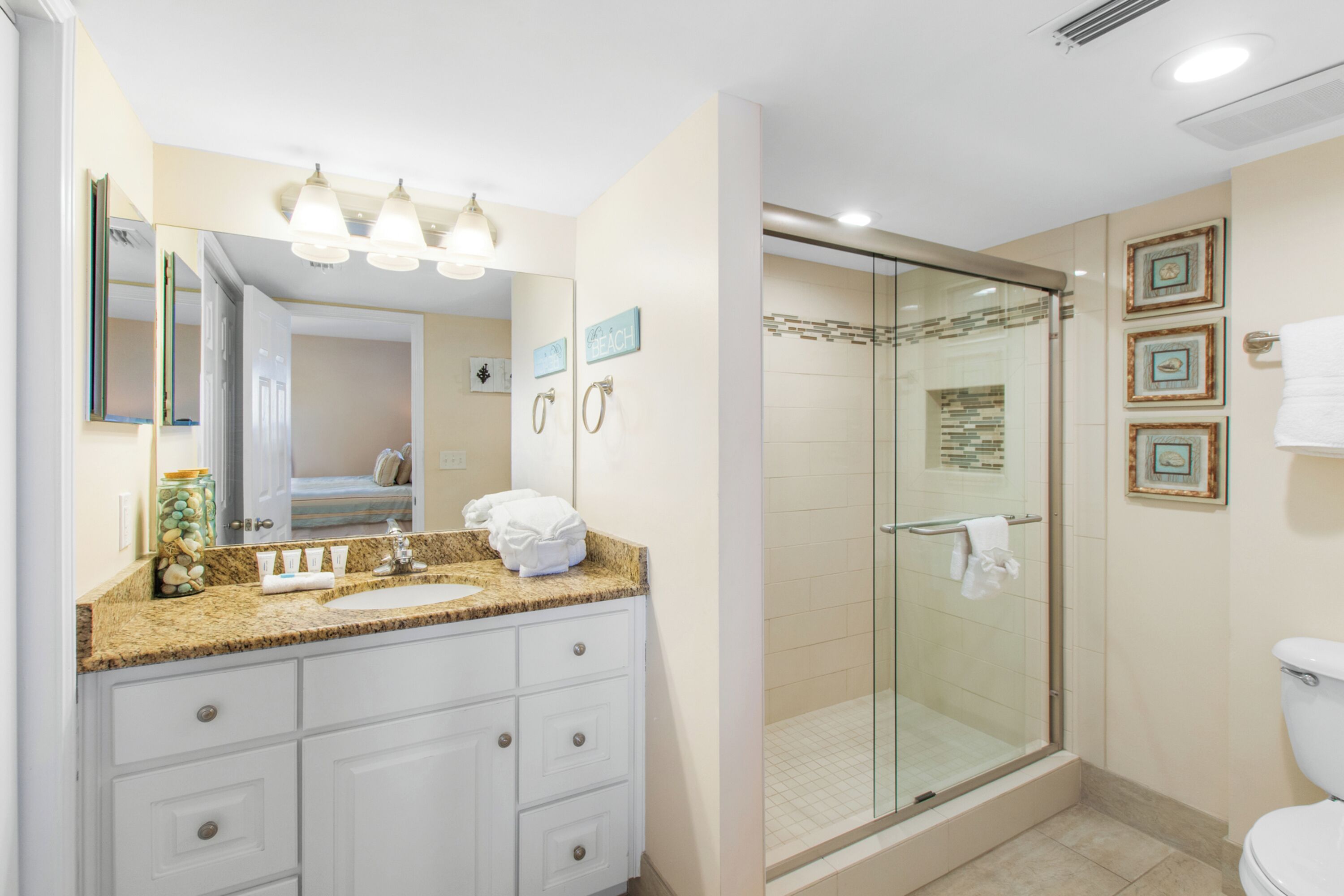 A modern bathroom with a granite countertop, white cabinetry, a glass-enclosed shower, and framed artwork on the wall.
