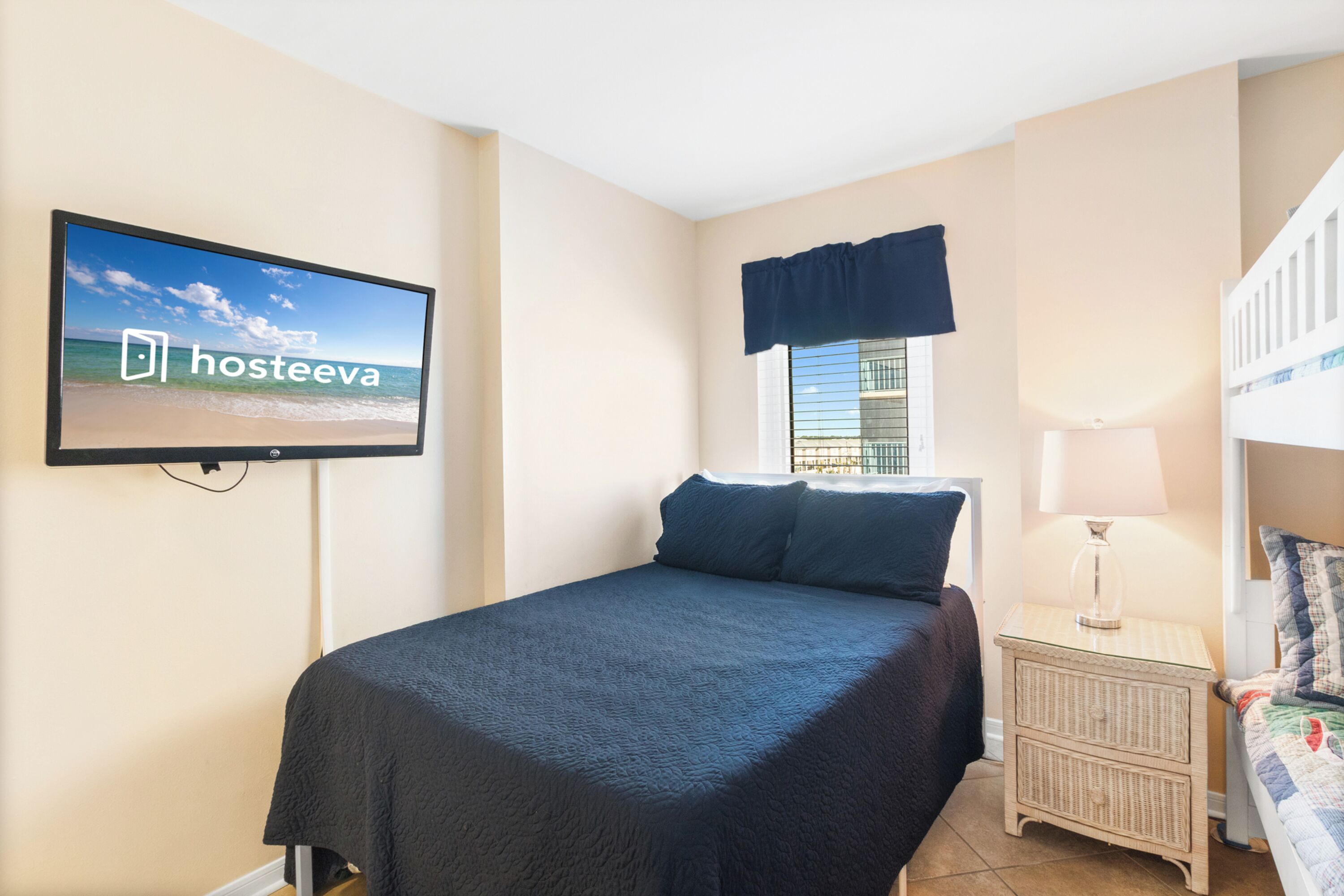 A bedroom with a bed, a nightstand, a TV displaying 'hosteeva', a window with a view, and a balcony.