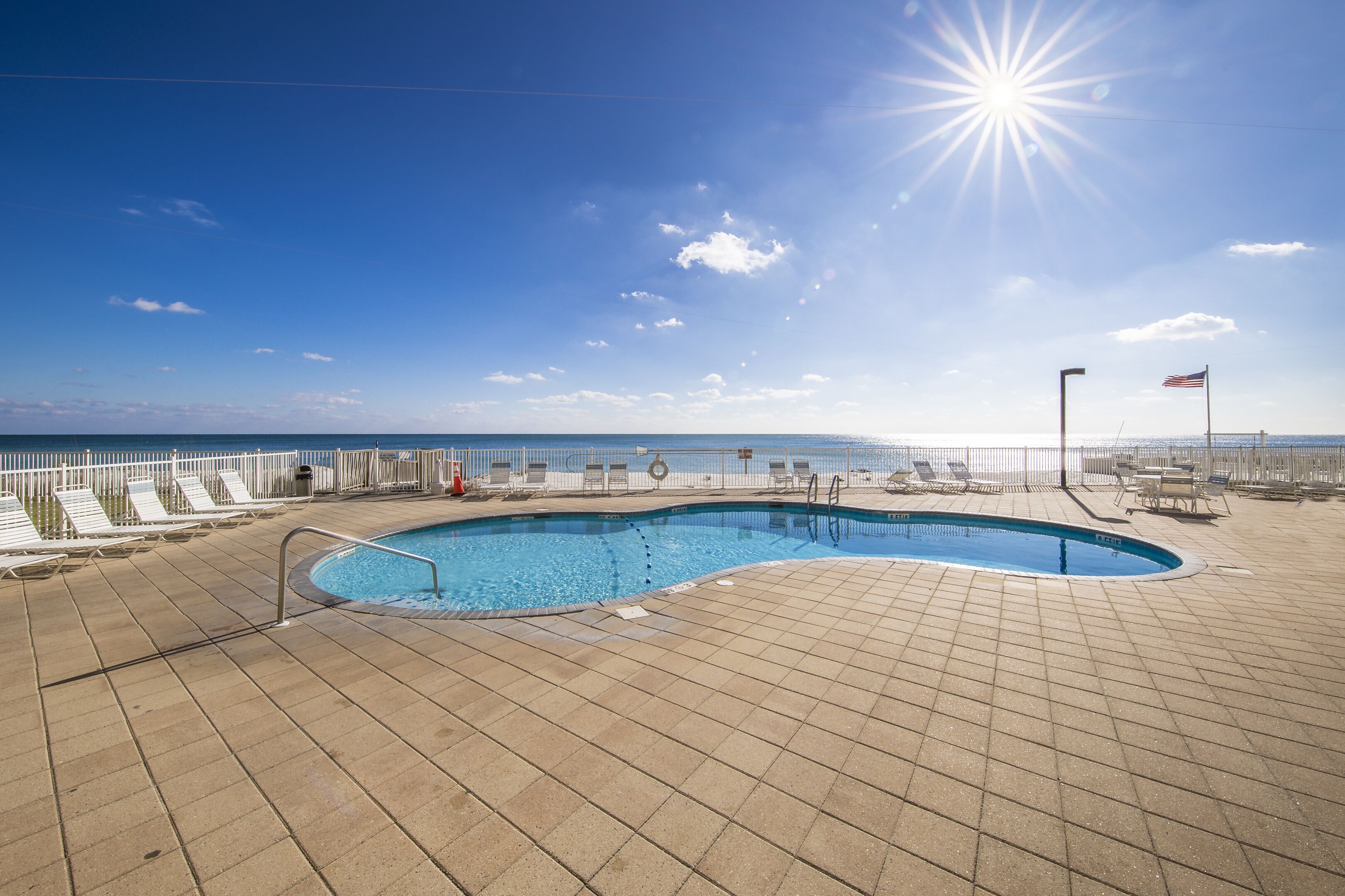 Una piscina iluminada por el sol con un borde de azulejos, sillas de descanso y vistas al océano.