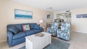 Een woonkamer met een blauwe bank, een witte salontafel en twee fauteuils met een patroon. Achtergrond toont een keuken.
