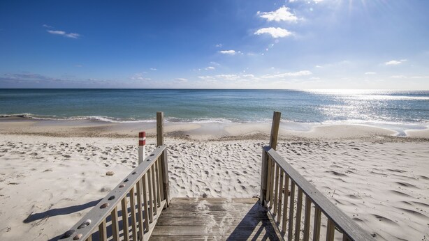 Een houten strandpromenade die naar de oceaan leidt, met een heldere lucht en schijnende zon.