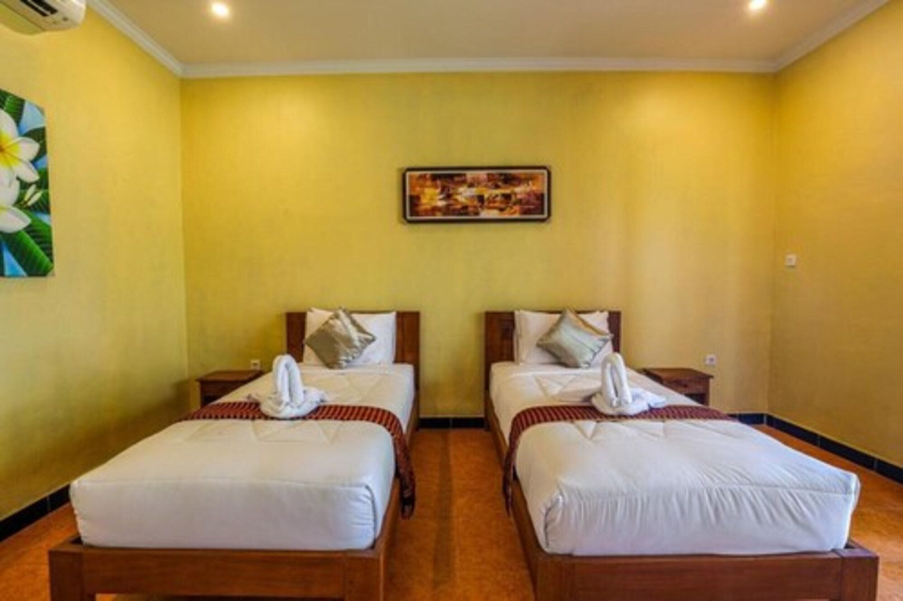 A hotel room with two single beds, a wooden headboard, a wall-mounted air conditioner, and a framed picture above the beds.