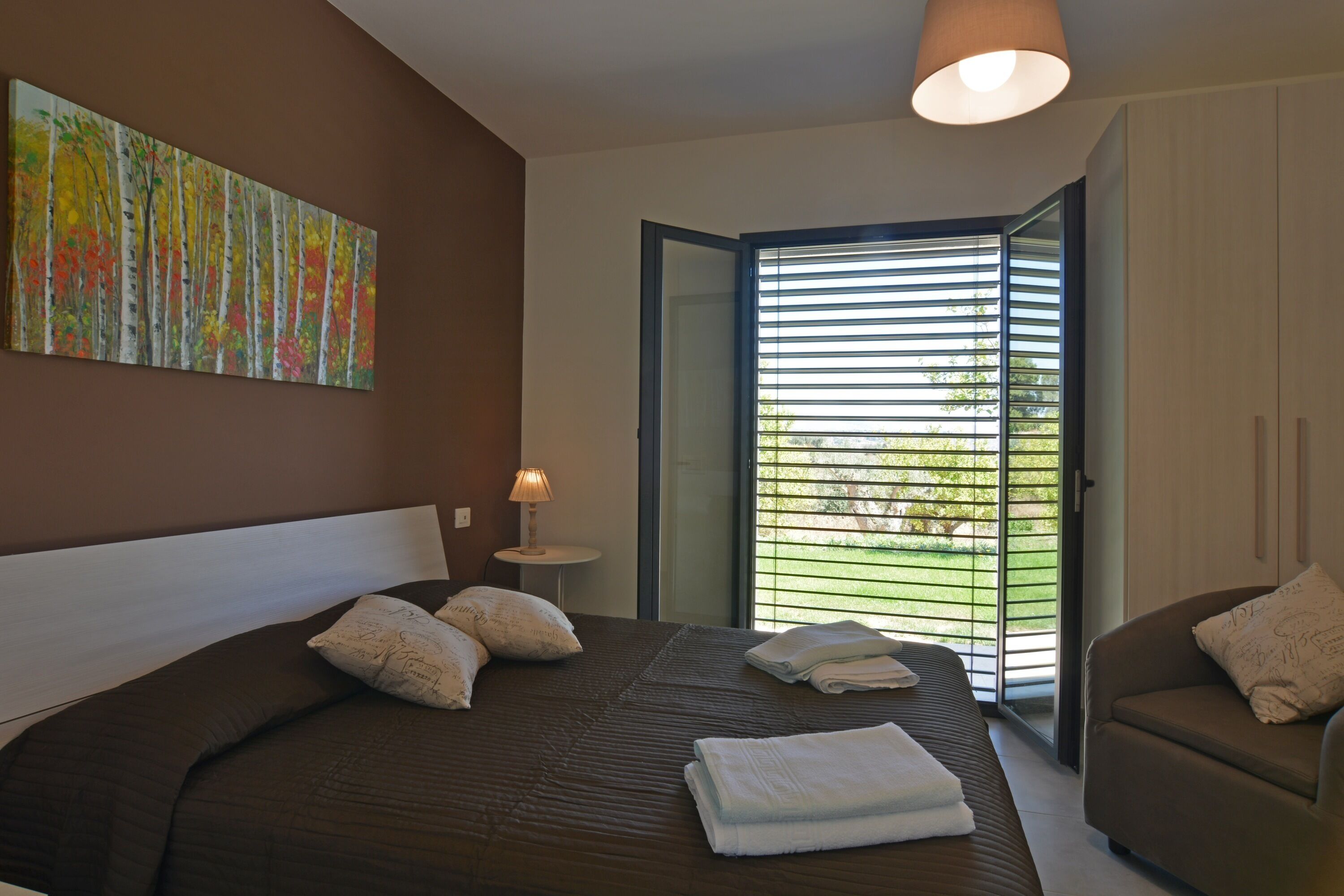 Une chambre avec un grand lit, une table de chevet avec une lampe, un canapé et une porte-fenêtre coulissante donnant sur un balcon.