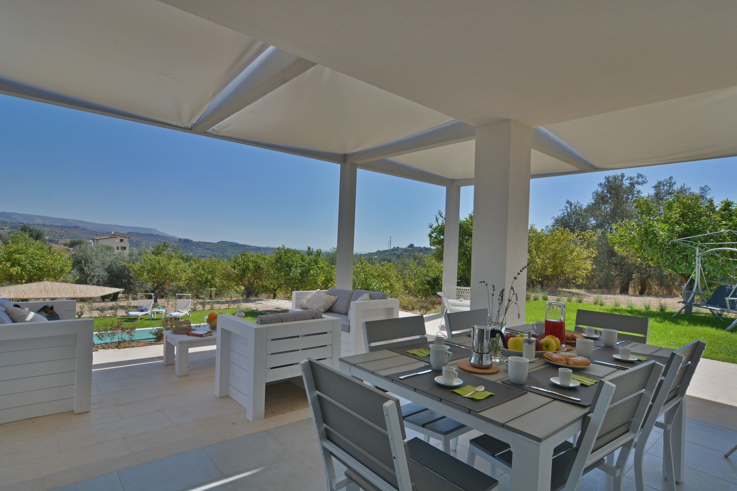 An outdoor patio with a table set for breakfast, white furniture, and a view of the countryside.