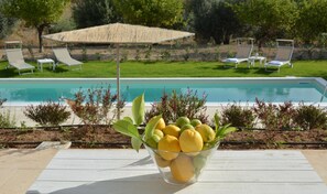 A poolside area with a glass bowl of lemons and limes, lounge chairs, and a thatched umbrella. - Villa Mennula in Noto (Noto)