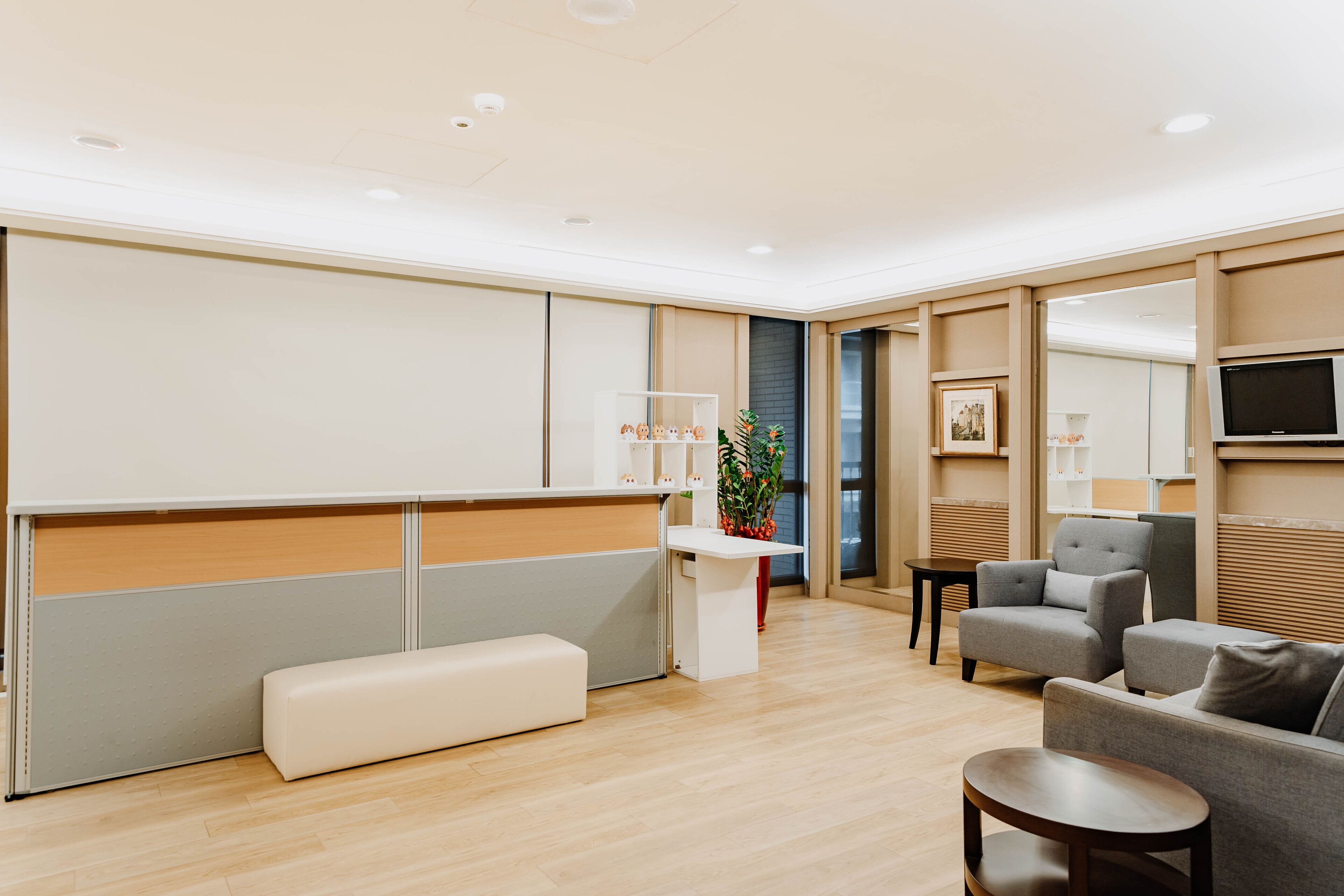 Een moderne hotelkamer met een houten vloer, een wit receptiebalie, een grijze bank, een klein rond tafeltje en een potplant.