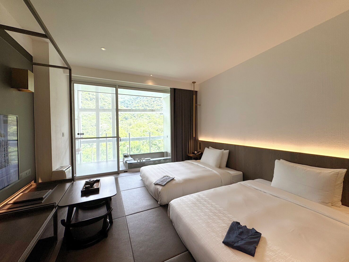 A hotel room with two beds, a desk, and a large window offering a view of greenery.
