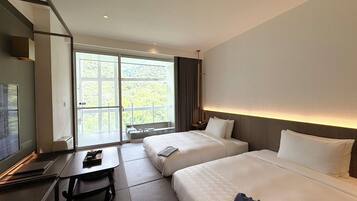 A hotel room with two beds, a desk, and a large window offering a view of greenery.