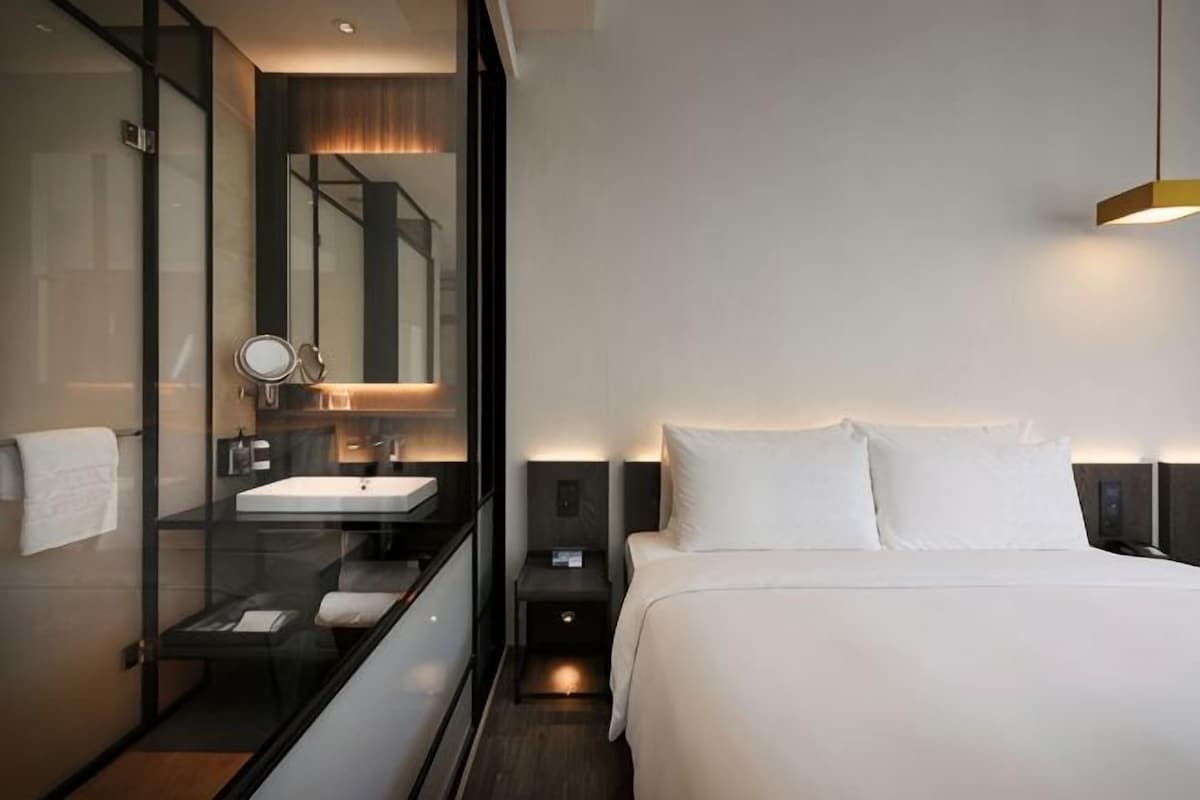 Ein modernes Hotelzimmer mit einem verglasten Badezimmer, einem Einzelbett, Nachttischen und einem Beistelltisch.