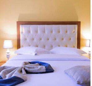 Una cama bien hecha con cabecera capitonada, ropa de cama blanca y una manta azul y gris.