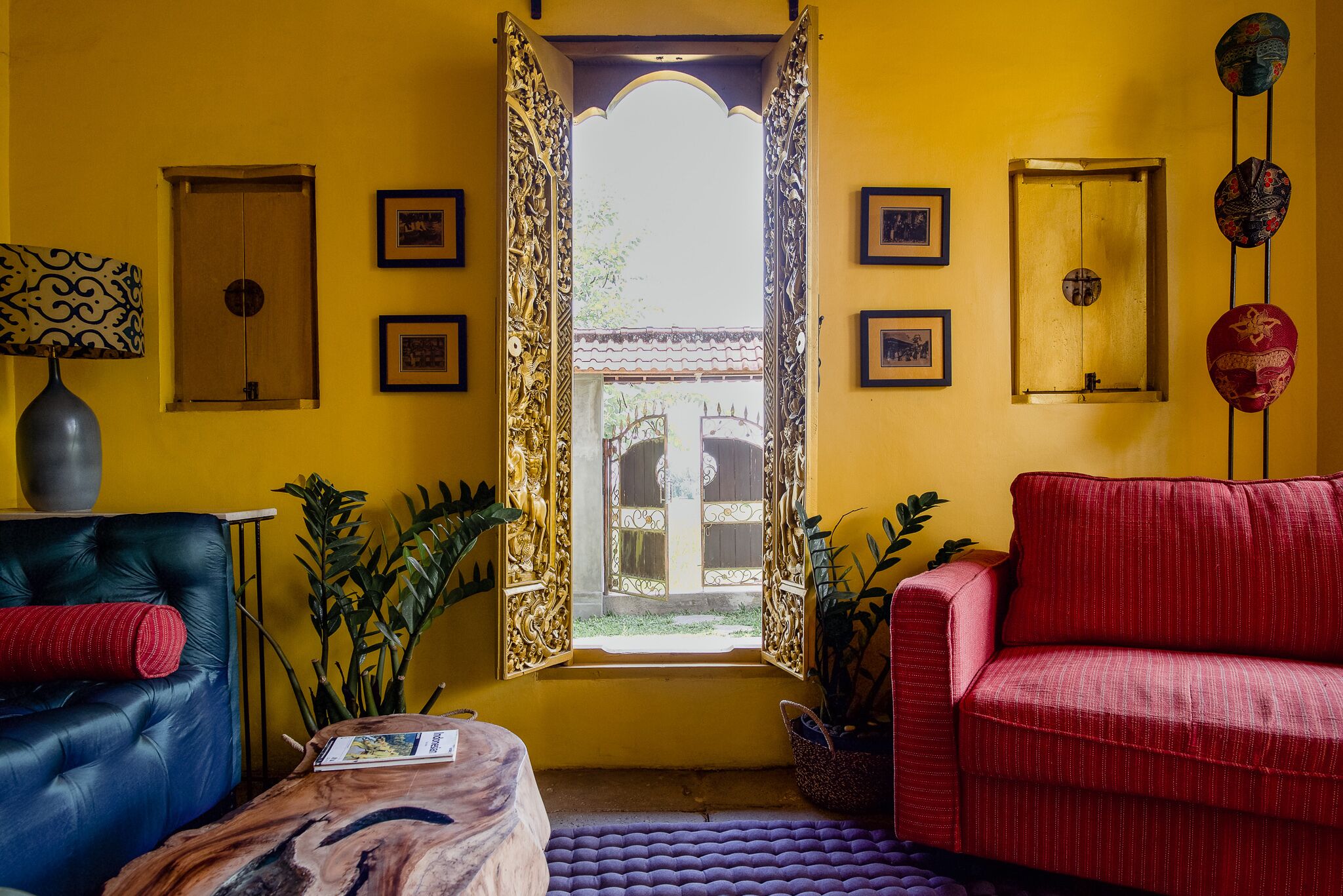 A room with a yellow wall, a red sofa, a blue sofa, and a wooden coffee table. There is a window with an ornate frame and a view of a courtyard.