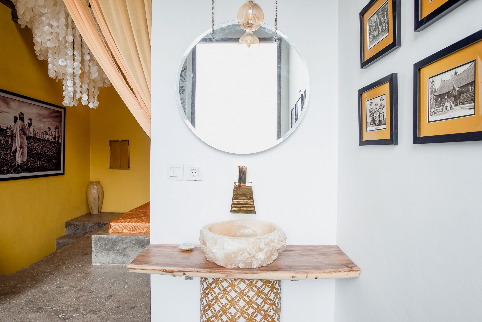 A bathroom with a round mirror, a wooden vanity, a gold faucet, and framed pictures on the wall.