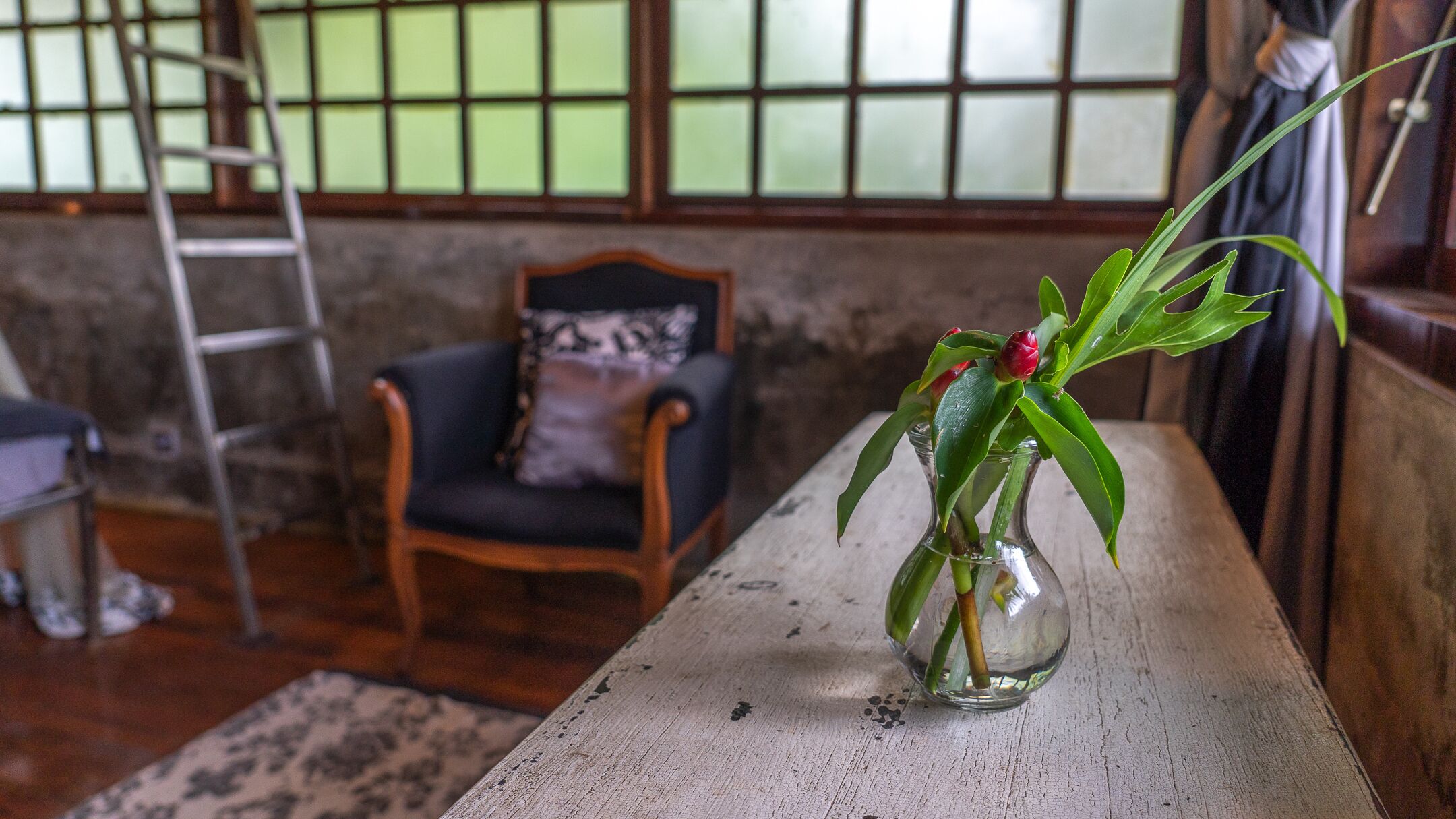 A room with a wooden table, a chair, a ladder, and a window with a view of the outside.