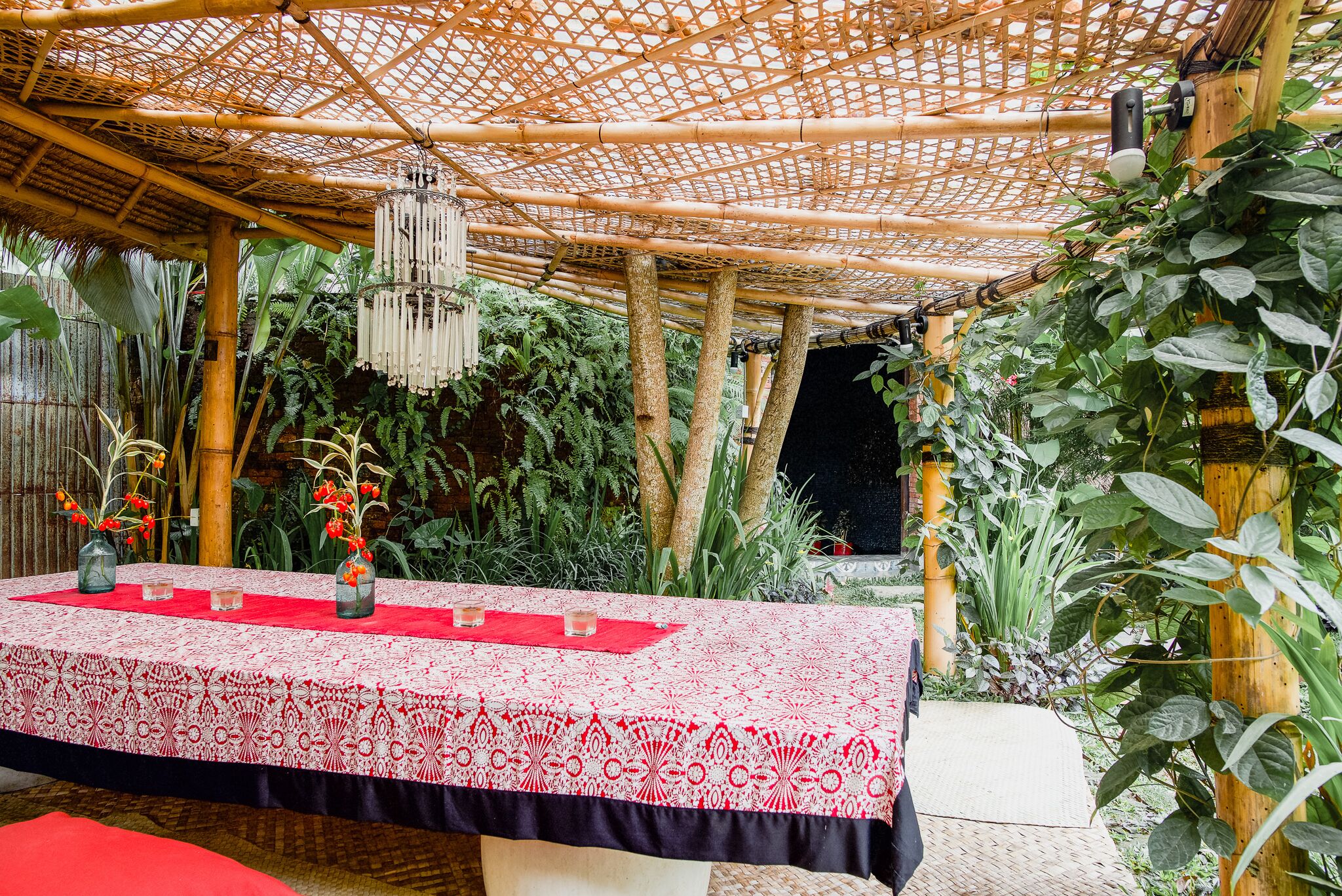 Un espace de restauration en plein air agrémenté d’une pergola en bambou, d’une table recouverte d’une nappe à motifs et de diverses plantes.