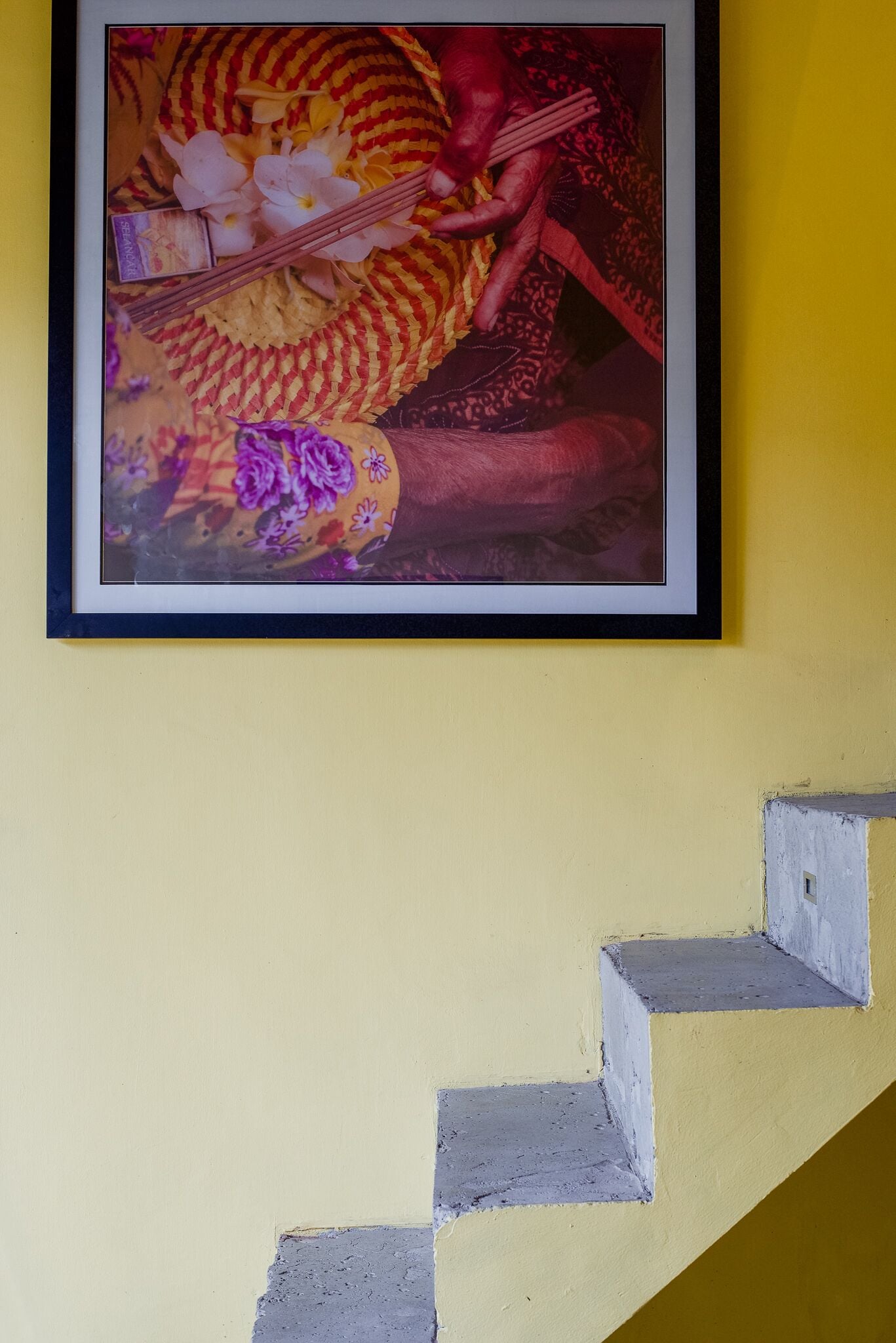 A framed artwork with a floral design and a pair of chopsticks on a yellow wall.