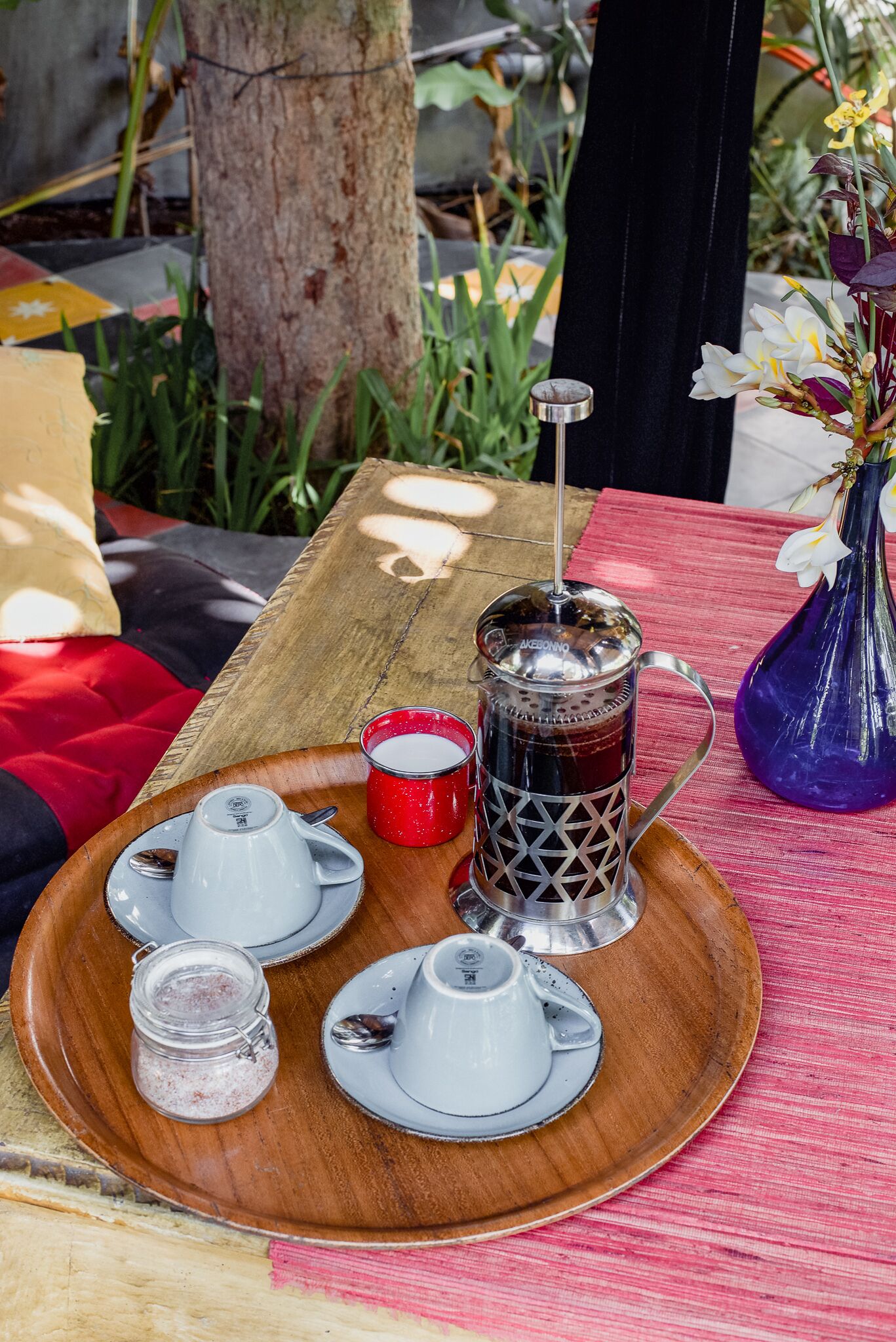 A wooden table with a coffee press, cups, and a small jar of sugar.