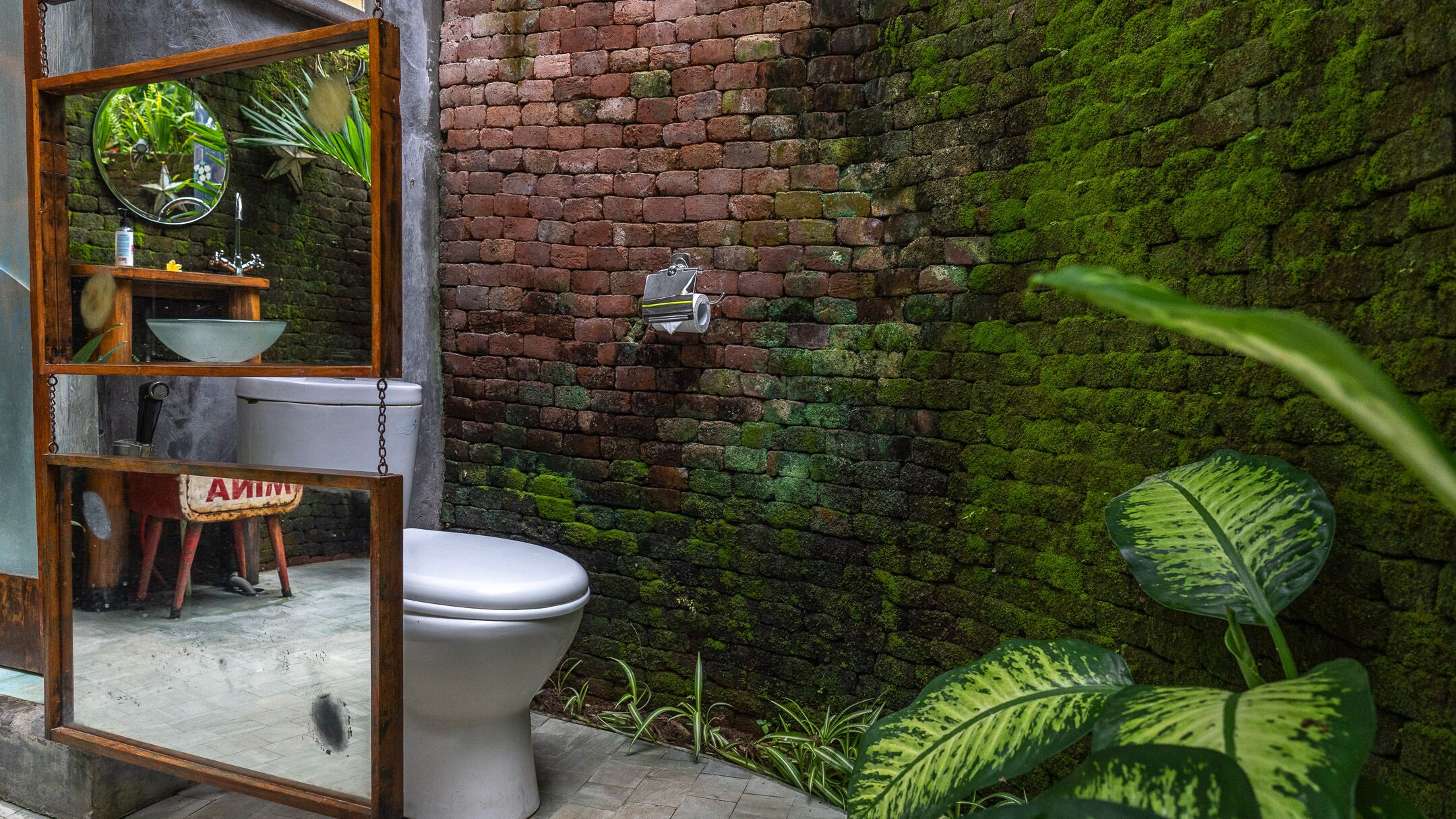 Un baño rústico con pared de ladrillo, inodoro blanco, estante de madera con espejo y plantas verdes.