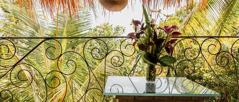 Una mesa con cubierta de vidrio, con un jarrón de flores, rodeada por una cerca de hierro forjado y palmeras.