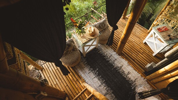 Habitación con suelo de bambú y vista a exuberante vegetación, una pequeña mesa con un sombrero y un farol, y una mesa blanca con varios objetos.