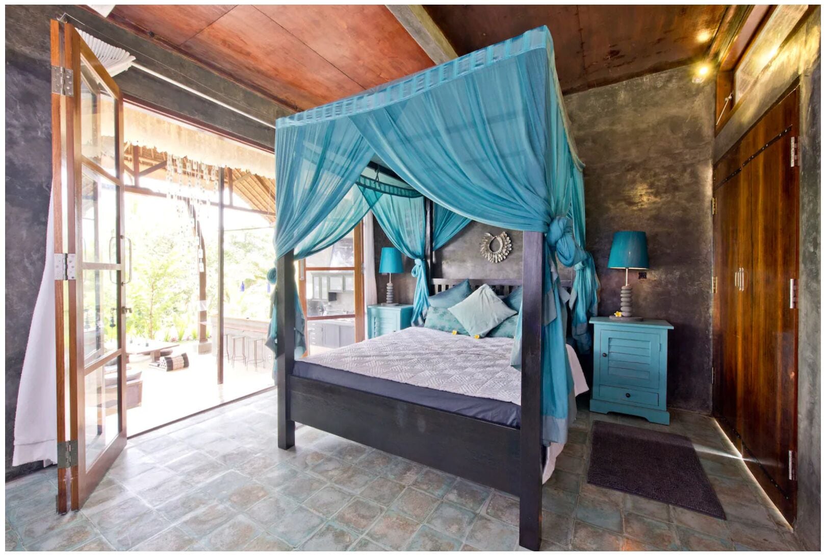 A four-poster bed with a canopy, a nightstand with a lamp, and a view of the outdoors.
