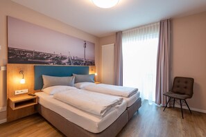 Two single beds and a comfortable chair await in this stylish room, featuring a large cityscape photograph and a convenient nightstand with a lamp and power outlet.