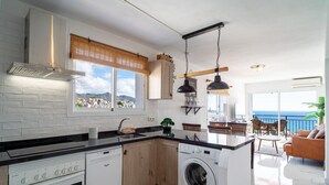 A modern kitchen with a built-in oven, dishwasher, and washing machine. It features a large window with a view of the sea and a balcony with outdoor seating.