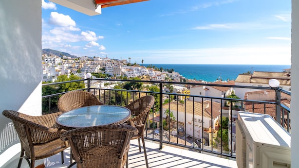 A balcony with wicker furniture and a glass table overlooking a coastal town.