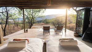 A bedroom with two beds, a wooden deck, a glass door, and a view of a mountainous landscape. - Sasi Bush Lodge (Jagersrust)