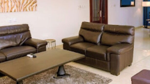 A living room with two brown leather sofas, a coffee table, and a patterned wall hanging. - Loumia Residency 3ch (Douala)