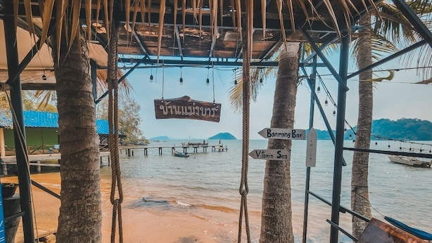 A wooden swing chair under a thatched roof, overlooking a beach with a pier and boats. Directional signs point to 'Dahiriang Bar' and 'Vilan Se'. - Banmae Resort Kohchang (Ko Chang)