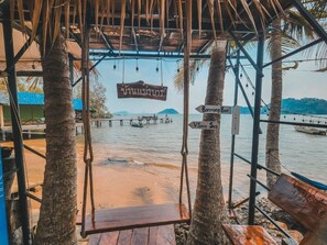 A wooden swing chair under a thatched roof, overlooking a beach with a pier and boats. Directional signs point to 'Dahiriang Bar' and 'Vilan Se'. - Banmae Resort Kohchang (Ko Chang)