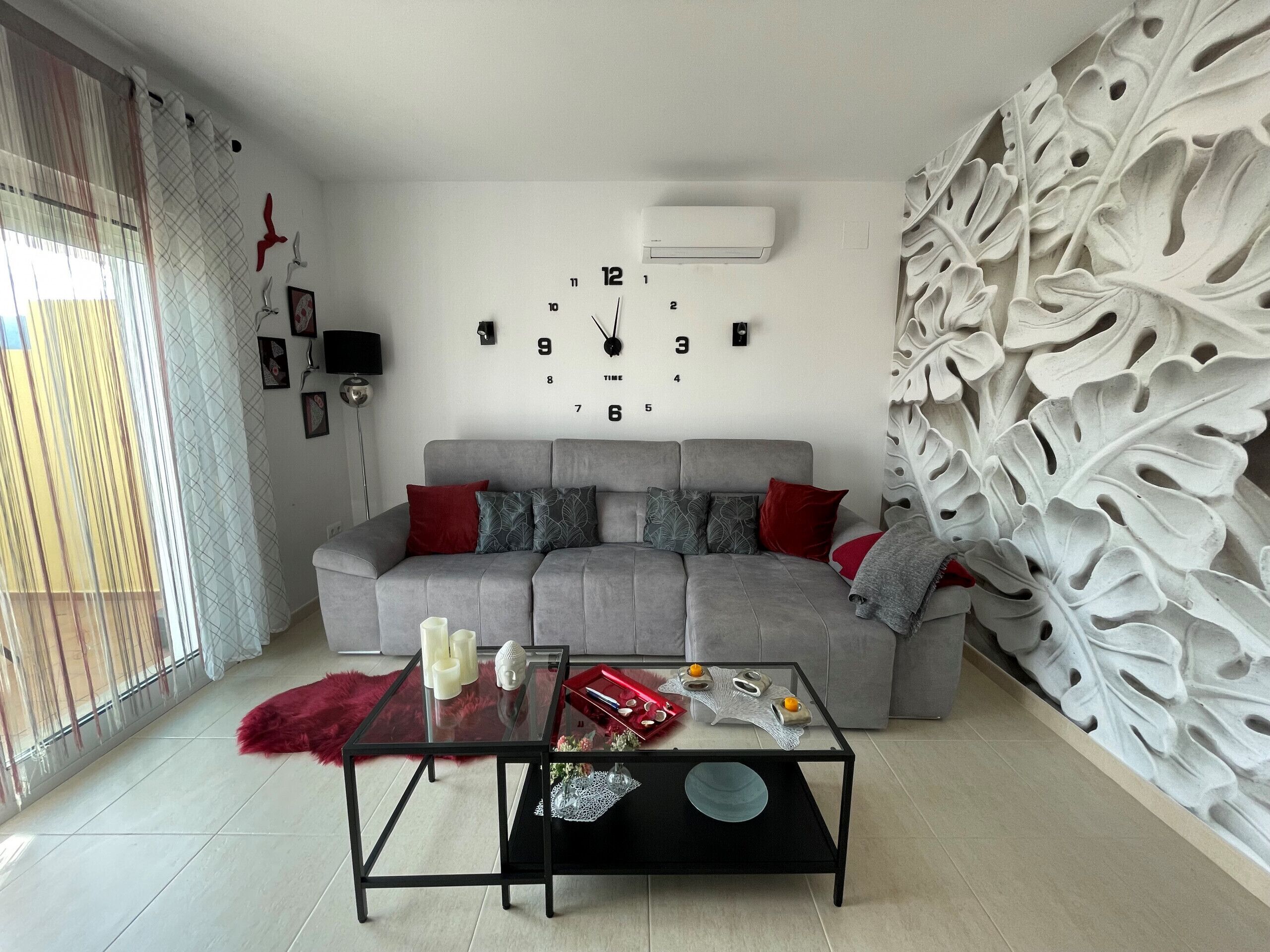 A modern living room with a grey sofa, red cushions, a glass coffee table, and a decorative wall with leaf patterns.