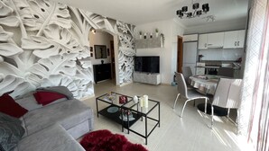 A modern living room with a grey sofa, a red furry rug, and a coffee table with decorative items. A flat-screen TV is mounted on the wall, and a dining area with a table and chairs is visible. - Green I - 4A (Pedreguer)