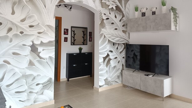 A modern living room with a large TV, a black entertainment unit, and a decorative wall with leaf patterns. - Green I - 4A (Pedreguer)