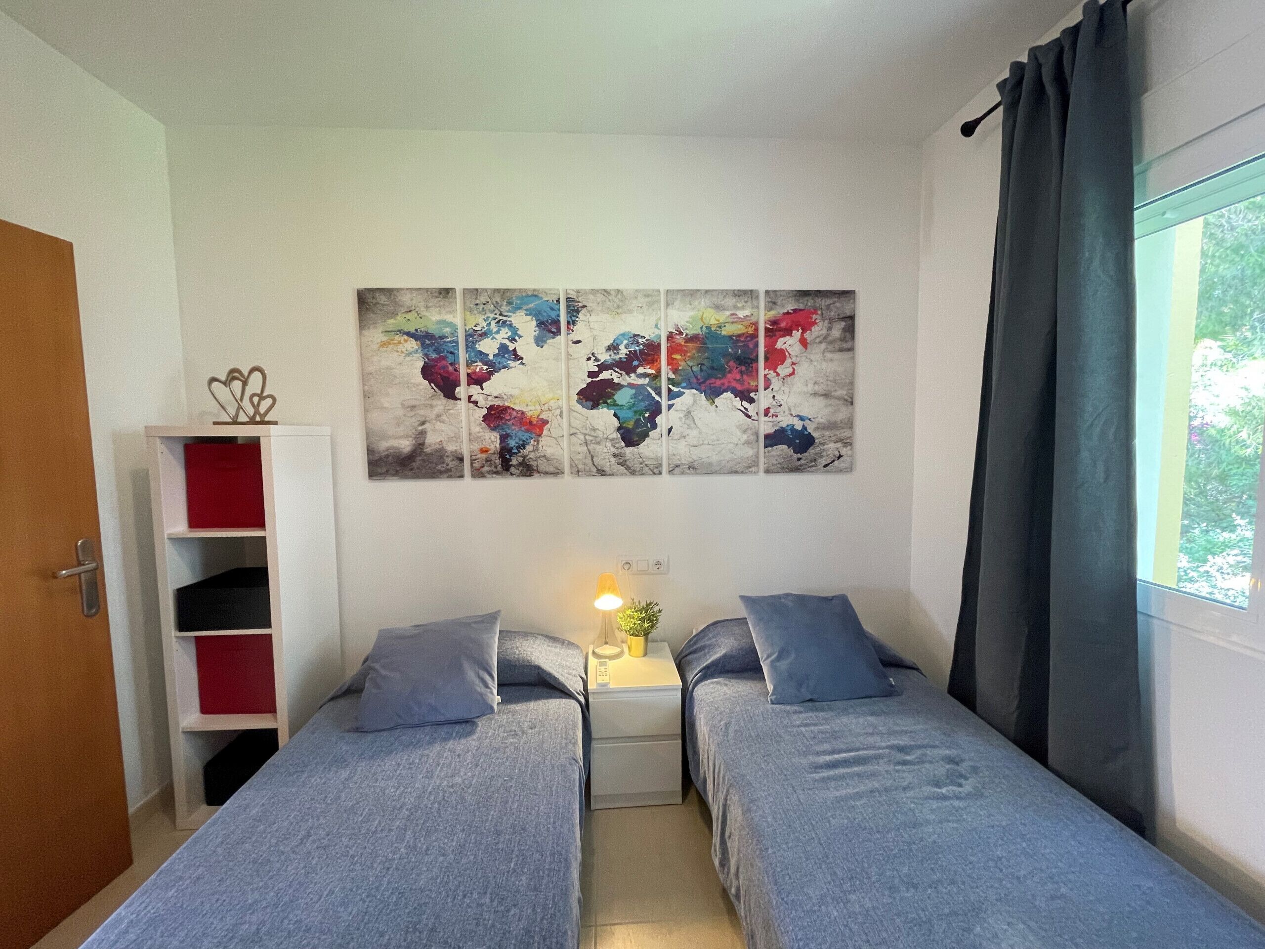 Une chambre avec deux lits simples, une table de chevet avec une lampe, une décoration murale représentant une carte du monde, une bibliothèque et une fenêtre avec des rideaux.