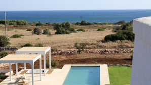 Zona de piscina con sillones de descanso y una pérgola, con vistas a un paisaje costero.