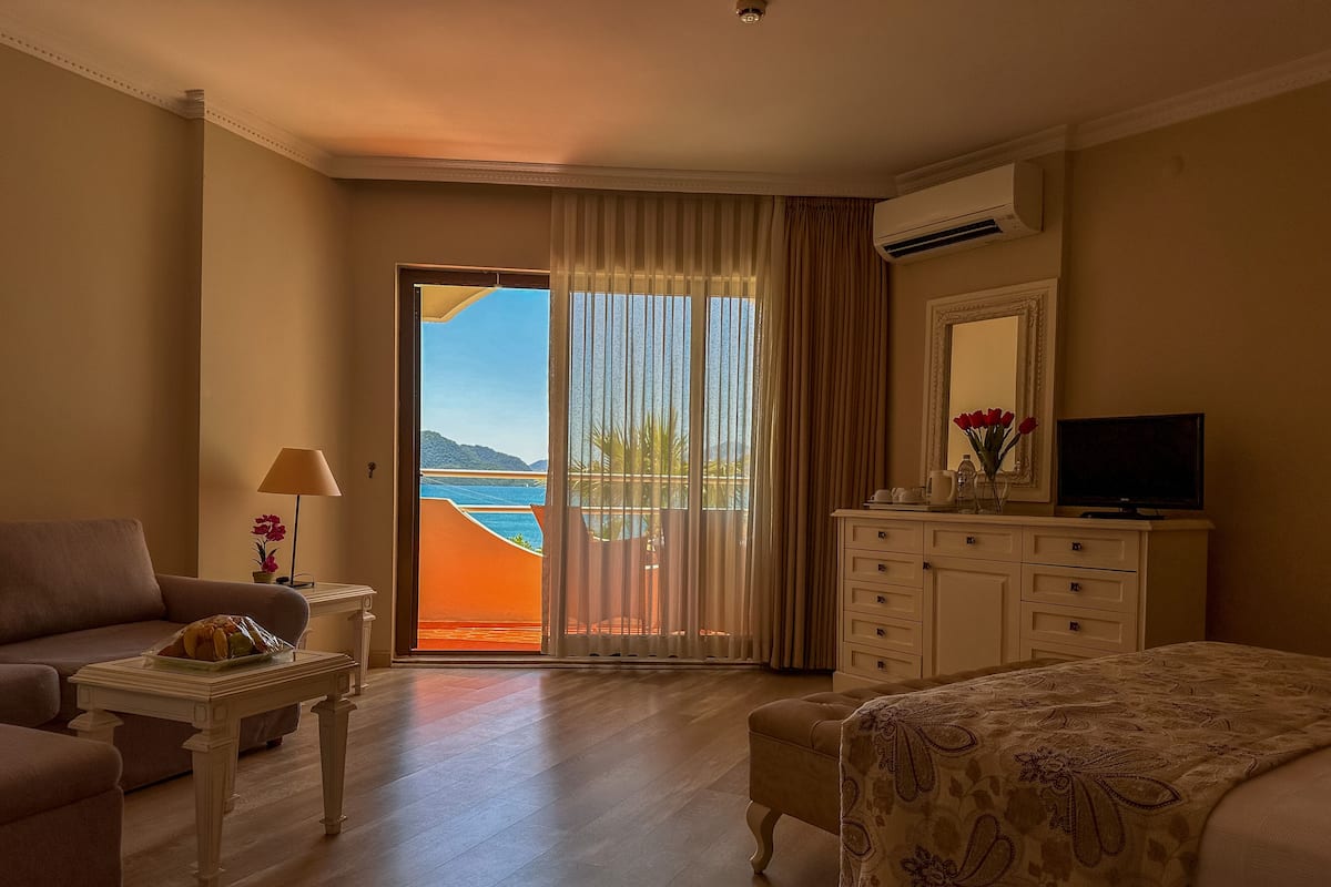 A hotel room with a bed, a TV, a dresser, a sofa, a table, and a view of the sea through a sliding door.
