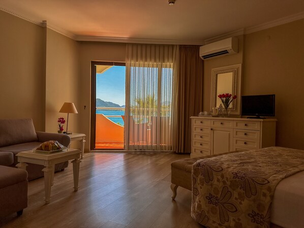 A hotel room with a bed, a TV, a dresser, a sofa, a table, and a view of the sea through a sliding door. - Hawaii Hotel (Marmaris)