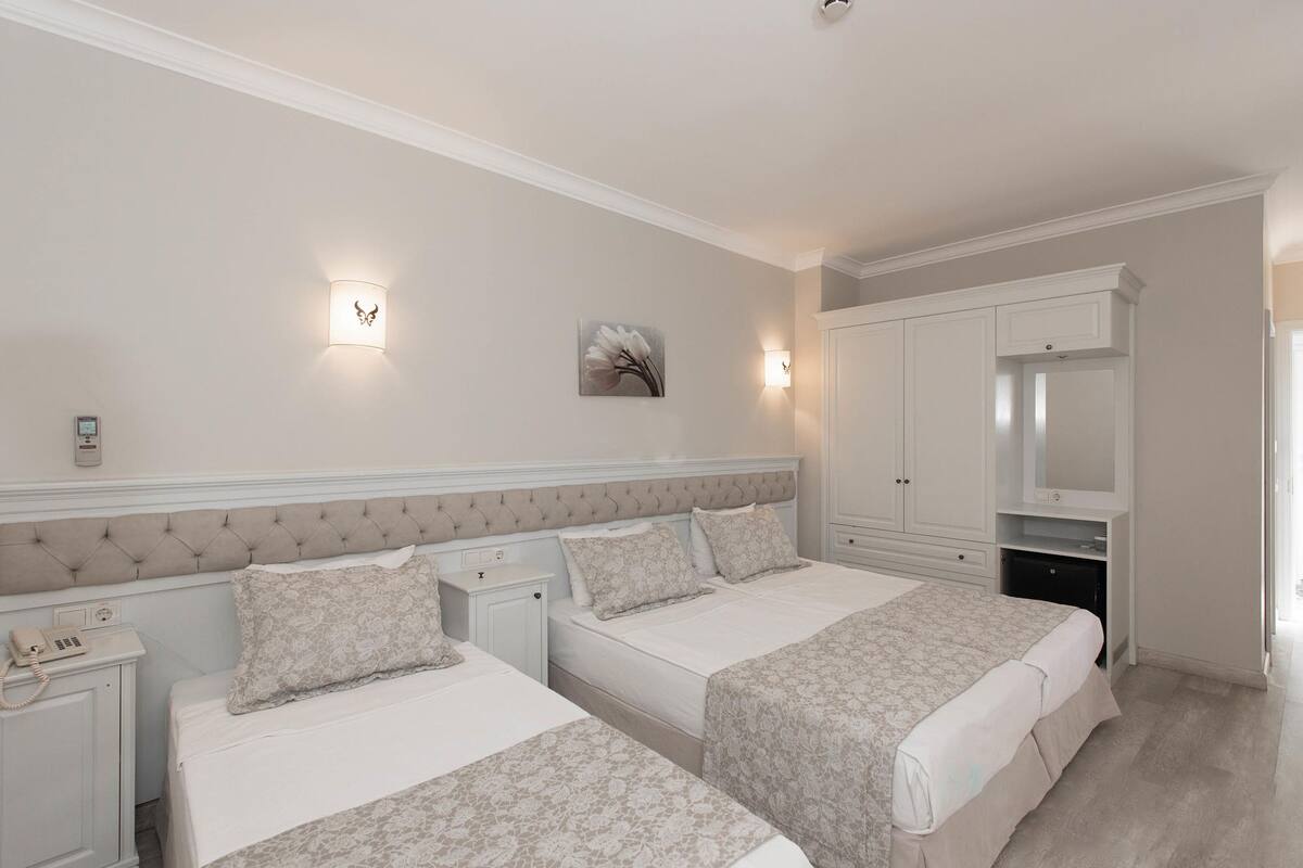 A hotel room with two beds, a wardrobe, a bedside table with a lamp, and a wall-mounted phone.