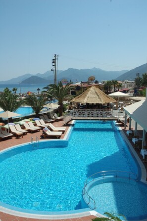 A swimming pool with lounge chairs and a thatched-roof structure, surrounded by palm trees and mountains in the background. - Hawaii Hotel (Marmaris)