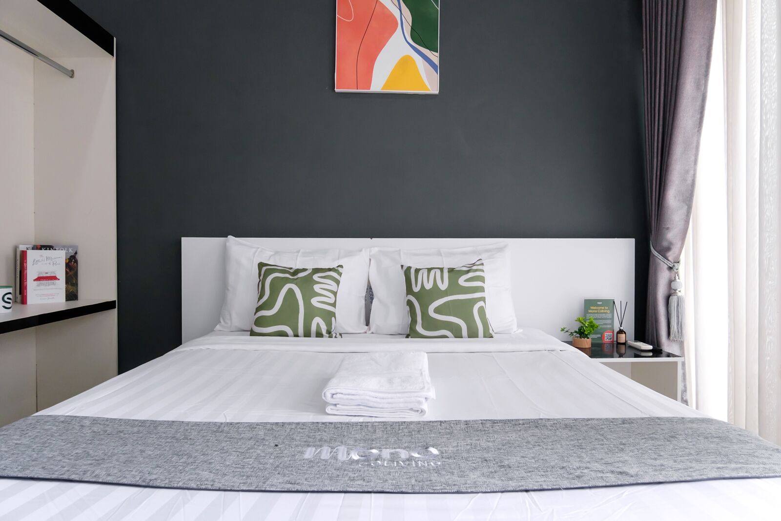 Een net opgemaakt bed met witte lakens, twee kussens met groene patronen, een grijze plaid en een kunstwerk erboven.