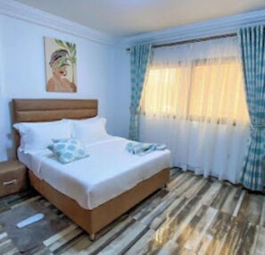 A bedroom with a wooden bed, white bedding, a nightstand, and a painting on the wall. - Belmann Bonapriso 1 ch (Douala)