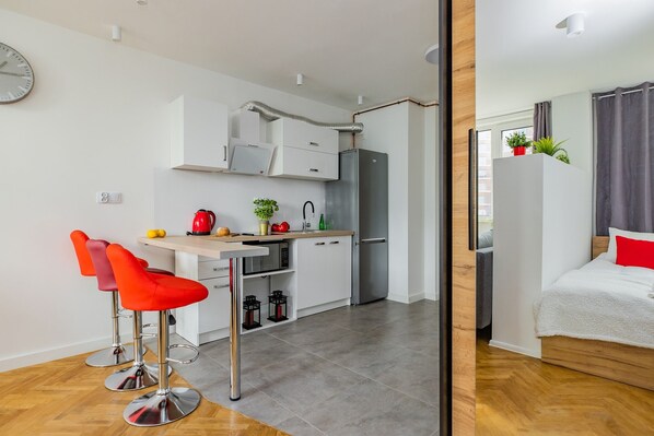 Een compacte keuken met moderne apparaten, een eethoek met rode stoelen en een slaapkamer met een bed en gordijnen.