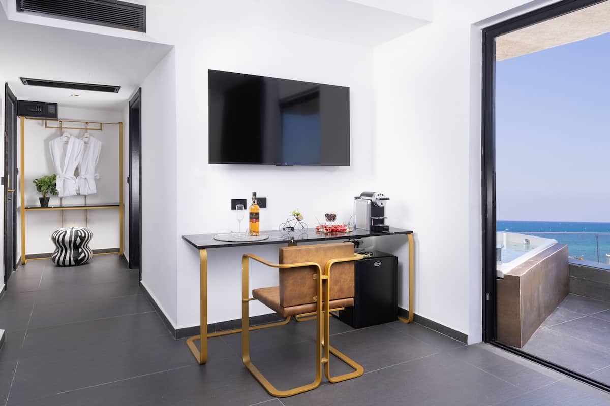 Una habitación de hotel moderna con balcón, televisor de pantalla plana, kitchenette y zona de estar con una silla y una mesa.