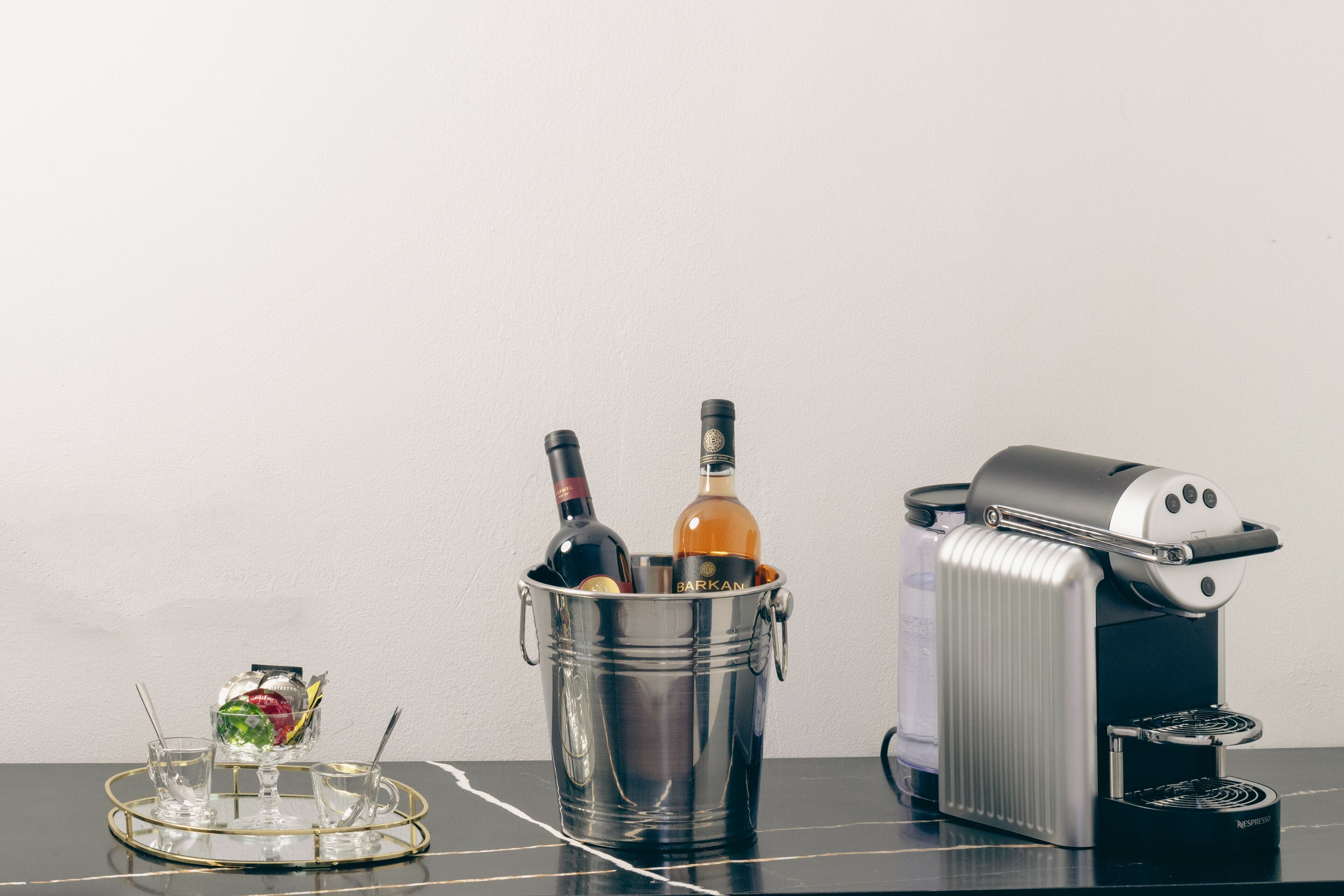 Un seau en acier inoxydable, deux bouteilles, un petit saladier en verre contenant des fruits et une machine à café Nespresso sont posés sur une table.