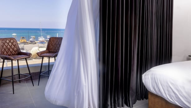Un balcón con dos sillas marrones, una cama con ropa de cama blanca y vistas al mar y a los barcos.