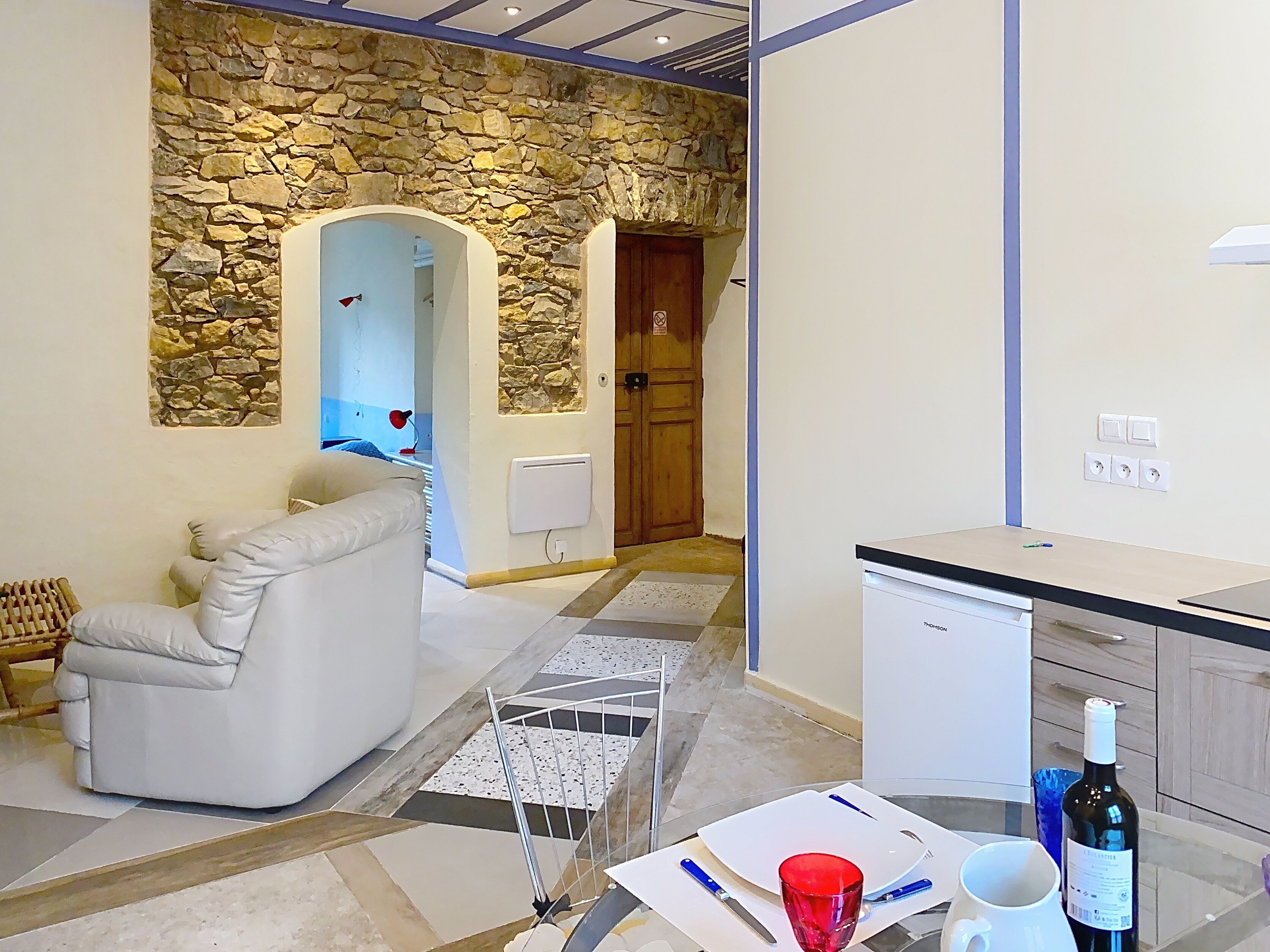 A modern living room with a stone wall, a white sofa, a dining area with a kitchen counter, and a red wine bottle.